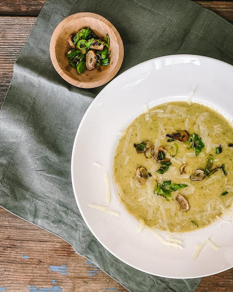 Leek and potato soup served in a white bowl with sauteed garlic chips and leeks tops on the side